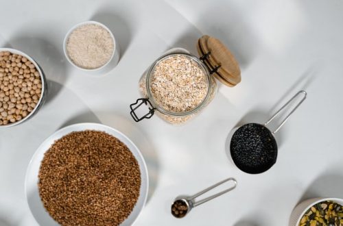 Top view of various grains and seeds in bowls and jars on a white surface.