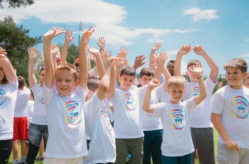 A group of cheerful children in white T-shirts raising hands in a park during summer.