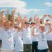 A group of cheerful children in white T-shirts raising hands in a park during summer.